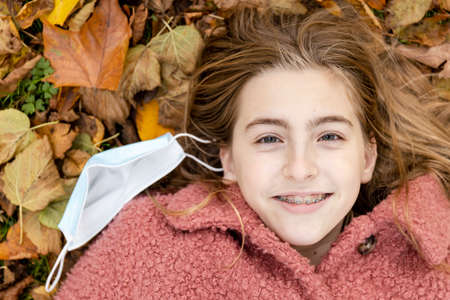 Teenage girl wearing braces smiling and laying in autumn leaves with a face mask besides her.の写真素材