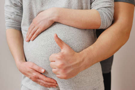 Hand of happy husband showing OK sign on background of stomach of wife on last month of pregnancyの写真素材