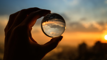 The hand holds glass ball which reflects sunset sky over city.の写真素材