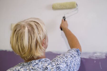 Authentic photo of aged woman painting interior wall with white paint.の写真素材