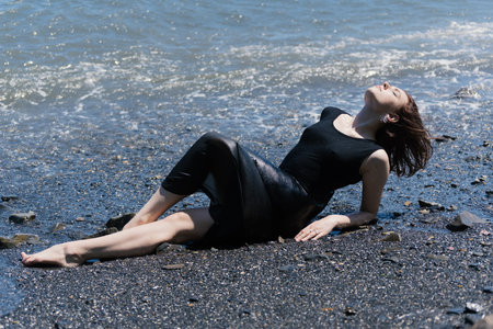 Young woman in black dress sitting on sand and enjoys sea.の写真素材