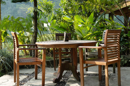 Empty wooden table and chairs are placed on a patio amidst lush tropical foliage in a garden.の写真素材