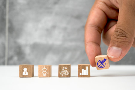A hand carefully places a wooden block with a target icon, symbolizing business goals, among other...