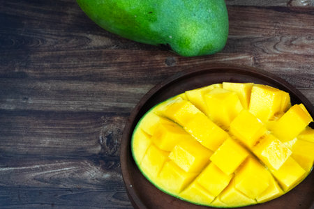 Sliced yellow mango half sits on a small wooden plate next to a whole green mango.の写真素材