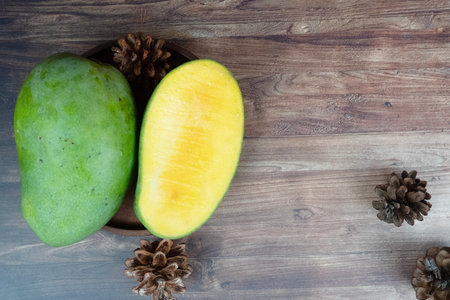 Green whole mango and sliced yellow half lie on a wooden plate with pinecones nearby.の写真素材