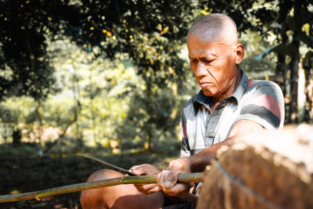 Elderly man carving bamboo with a knife in natural outdoor settingの写真素材