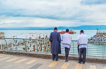 Three young Orthodox Hasid friends in traditional skullcaps and tallits stand on embankment of the Sea of Galilee and enjoy the view, Tiberias, Israel.のeditorial素材
