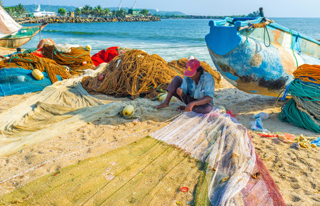 GALLE, SRI LANKA - DECEMBER 3, 2016: The fisherman sews the net, sitting next to the old catamaran boat on the sandy beach, on December 3 in Galle.のeditorial素材