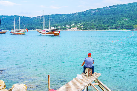 The fisherman on his pier enjoys his hobby and a view on ships in Dakapo harbor, Kemer, Turkey.のeditorial素材