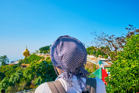 The tourist in headscarf watches the Golden Rock Shrine on the Kyaiktiyo mountain's edge from the viewpoint of the religious complex, Myanmar.の写真素材