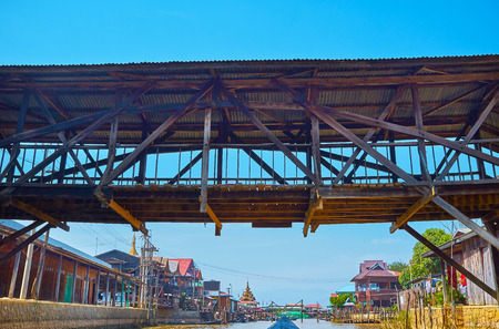 Floating under the old wooden bridge above the narrow canal of Inle Lake with dense housing on the...