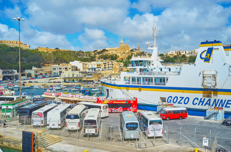 GHAJNSIELEM, MALTA - JUNE 15, 2018: Numerous tourist buses are parked at the ferry terminal of Gozo Island, the large ro-ro ferry and Lourdes Chapel are seen on the background, on June 15 in Ghajnsielem.のeditorial素材