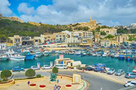 GHAJNSIELEM, MALTA - JUNE 15, 2018: Mgarr harbour with old fishing boats is surrounded by tall hills with residential cottages and Lourdes Chapel on the top, Gozo Island, on June 15 in Ghajnsielem.のeditorial素材
