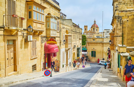 VICTORIA, MALTA - JUNE 15, 2018: The medieval cityscape with stone edifices, tourist stores, side wall of Local Council and the huge dome of St George Basilica on the background, on June 15 in Victoria.のeditorial素材
