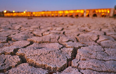 The close-up of cracked mud on bottom of Zayanderud river with a view on Siosepol bridge in bright evening lights on the background, Isfahan, Iran.のeditorial素材