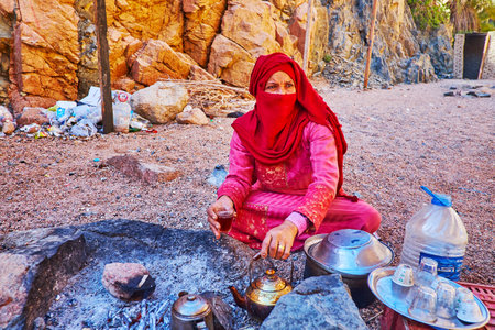 DAHAB, EGYPT - DECEMBER 16, 2017: The Bedouin  woman in bright red niqab makes hot tea, boiled on charcoal in desertのeditorial素材