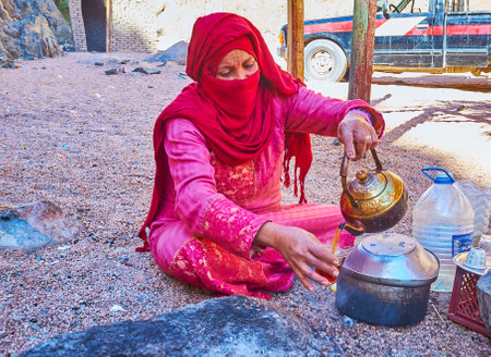 DAHAB, EGYPT - DECEMBER 16, 2017: The portrait of Bedouin woman in niqab, she, sits on the sand and pours black tea to the glassのeditorial素材