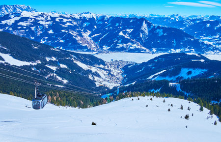 The beautiful winter landscape of Alps from the peak of Schmittenhohe mount, overlooking its snowy slope with ski trails, riding modern air lift of Schmittenhohebahn cableway and frozen Zeller see lake, Zell am See, Austriaのeditorial素材