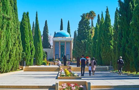 SHIRAZ, IRAN - OCTOBER 14, 2017: People walk the cypress alley of Tomb Saadi (Mausoleum of Sa'di, Sadiyeh) with flower bed amid the way and richly decorated pavilion on background, on October 14 in Shirazのeditorial素材