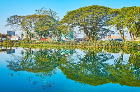 Idyllic nature of Nyaungshwe tourist village; the line of tall shady trees is reflected on mirror surface of Tharzi pond, Myanmarの写真素材