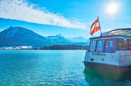 ST WOLFGANG, AUSTRIA - FEBRUARY 23, 2019: Bright blue rippled surface of Wolfgangsee lake with waving Austrian flag on the ferry's stern and rocky Alps on opposite bank, on February 23 in St Wolfgangのeditorial素材