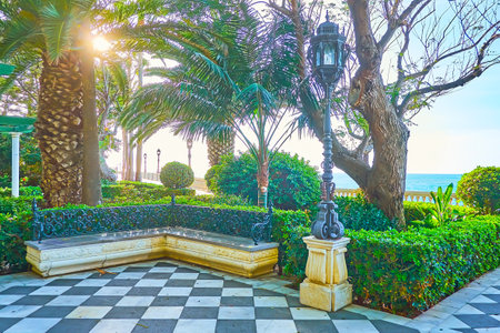 The evening sun shines through the lush palm tree of Paseo de Carlos III garden, decorated with retro streetlights, stone benches and topiary plants, Cadiz, Spainのeditorial素材