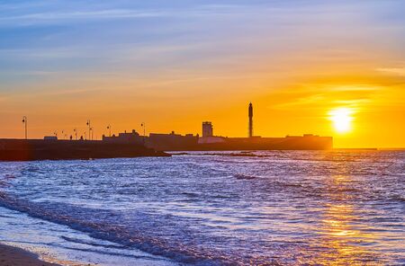 Enjoy the sunset over Atlantic Ocean with a view on tidal waves and silhouette of medieval San Sebastian castle with tall lighthouse, Cadiz, Spainの写真素材