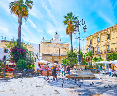 SANLUCAR, SPAIN - SEPTEMBER 22, 2019: People feed the pigeons in historic Plaza del Cabildo square with vintage lanterns, lush greenery and Municipal Library building on background, on September 22 in Sanlucarのeditorial素材
