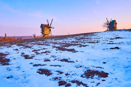 The snowy field in the early spring with bright sunset sky above it and the old timber windmills on horizon, Pyrohiv Skansen, Kyiv, Ukraineのeditorial素材