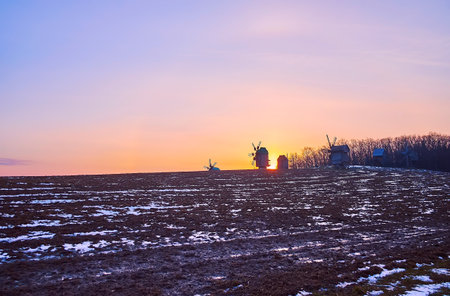 The evening view of the plowed snowy field, fiery sunset and the old timber windmills on the horizon, Pyrohiv Skansen, Kyiv, Ukraineのeditorial素材