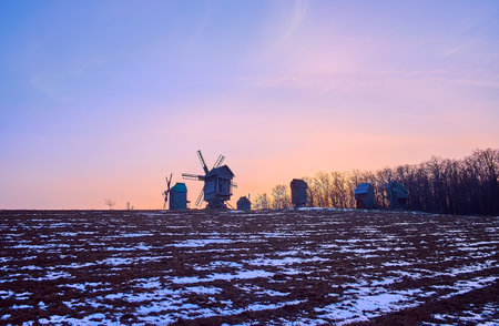 The bright dusk sky over the snowy plowed field and preserved historic timber windmills of Pyrohiv Skansen, Kyiv, Ukraineのeditorial素材