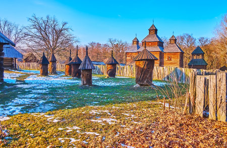 Watch traditional log hives in the old Polissya region farmstead with the wooden Resurrection church, seen behind the fence, Pyrohiv Skansen, Kyiv, Ukraineのeditorial素材