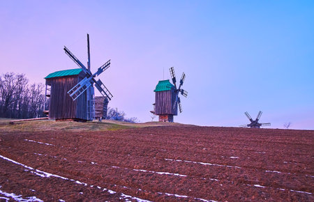 The beautiful wooden windmills of Pyrohiv Skansen with a plowed field in the foreground and bright purple dusk sky, Kyiv, Ukraineのeditorial素材