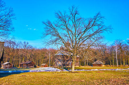 The timber houses, barns, historic Resurrection Church from Polissya Region are seen behind the green lawn of Pyrohiv Skansen, Kyiv, Ukraineのeditorial素材