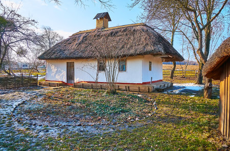 Traditional rural scene for Ukrainian village -whitewashed hata farmhouse with straw roof, surrounded with remains of the last spring snow in the wet swampy courtyard, Pyrohiv Skansen, Kyiv, Ukraineのeditorial素材