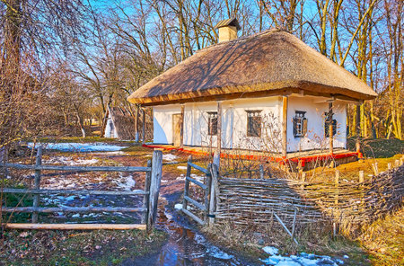 The scenic whitewashed hata house amid the snowy and swampy court, seen behind the rural wicker fence, Pyrohiv Skansen, Kyiv, Ukraineのeditorial素材