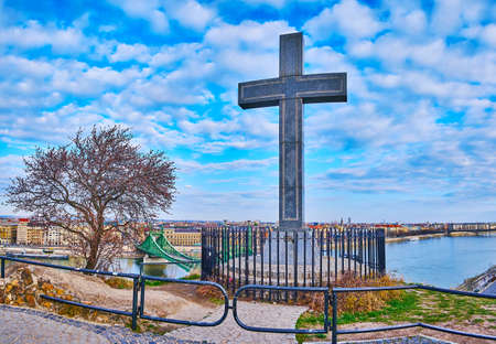 The stone Cross of Cave Church on the Gellert Hill against the Liberty Bridge and bright blue sky, Budapest, Hungaryの写真素材