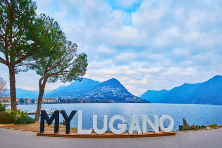 LUGANO, SWITZERLAND - MARCH 14, 2022: The black and white MyLugano sign on pedestrian embankment of Lake Lugano against Monte Bre at dusk, on March 14 in Luganoのeditorial素材