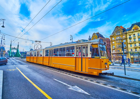 The vintage yellow tram at the stop on Fovam Square against the backdrop of historic townhouses and green Liberty Bridge, Budapest, Hungaryの写真素材