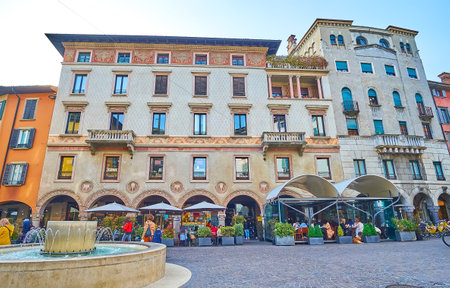 The frescoed facade of the historic edifice on Largo Nicolo Rezzara with outdoor dinings and the modern stone fountain in the foreground, Citta Bassa, Bergamo, Italyの写真素材