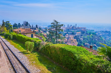 The houses, bell towers and churches of Citta Alta of Bergamo behind the trimmed bushes and rails of Bergamo-San Vigilio funicular, San Vigilio Hill, Bergamo, Lombardy, Italyの写真素材