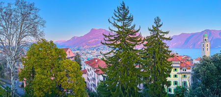 Evening Lugano panorama with Alps, Lake Lugano, bell tower of an Lorenzo Cathedral and tall trees in the foreground, Ticino, Switzerlandの写真素材