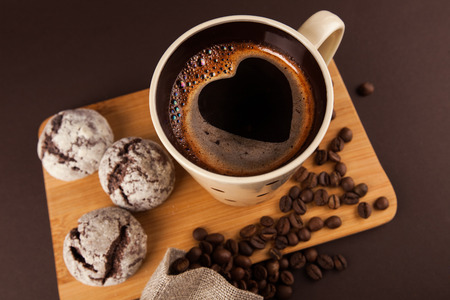 Cup of coffee with foam, heart shaped, with cookies and coffee beans, lying on the wooden stand, on brown background