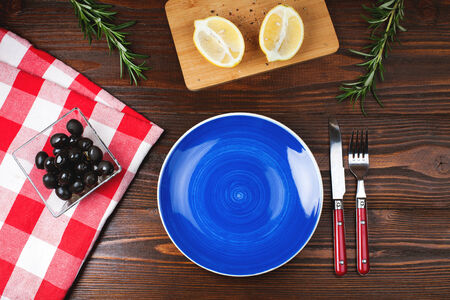 Blue plate with fork and knife, on  wooden table with olives, lemon and rosemaryの写真素材