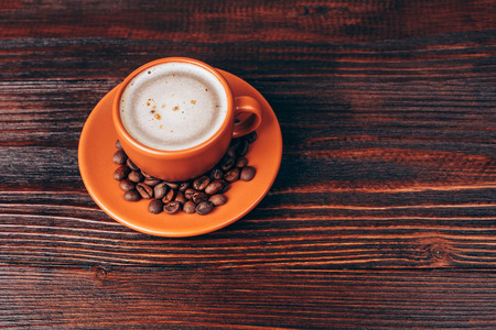 Ceramic orange cup of coffee with foam and coffee beans, standing on wooden tableの写真素材