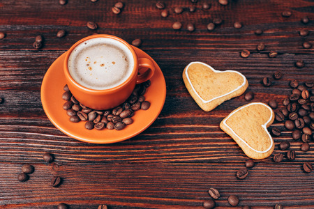 Ceramic orange cup of coffee with foam, with heart shaped cookies and coffee beans, lying on wooden tableの写真素材