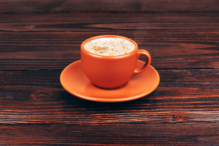 Ceramic orange cup of coffee with foam, standing on wooden table on wooden backgroundの写真素材
