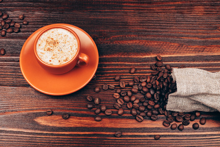 Ceramic orange cup of coffee with foam and coffee beans, standing on wooden tableの写真素材