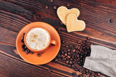 Ceramic orange cup of coffee with foam, with heart shaped cookies and coffee beans, standing on wooden tableの写真素材