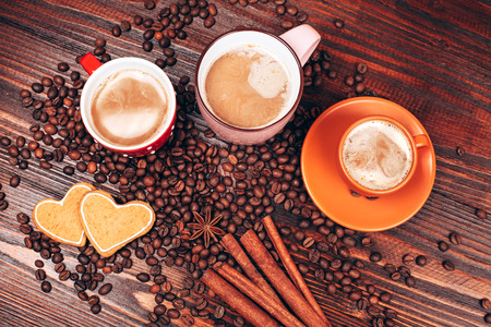 View from above of closely packed mugs full of hot coffee with foam, cinnamon sticks, star anise, coffee beans and wooden board with heart shaped cookies, staying on the wooden tableの写真素材